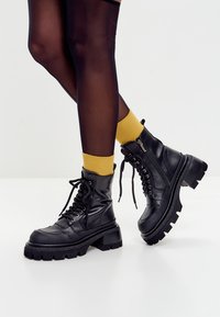 Gambe in collant neri trasparenti, calzini giallo senape, indossando stivaletti neri con piattaforma spessa e lacci, con cerniere laterali su un pavimento bianco.