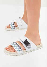Hvide sandaler med geometriske mønstrede stropper i sort, turkis og grå; har justerbare spænder og strukturerede gummisåler.