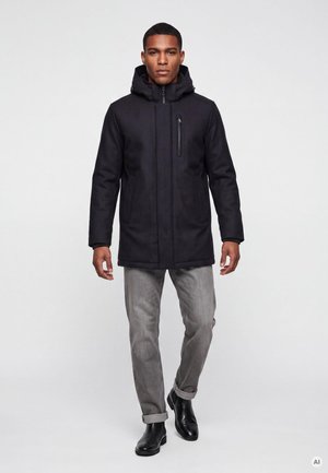Mann trägt einen schwarzen Wintermantel mit Kapuze, graue Jeans mit hochgekrempeltem Saum und schwarze Lederstiefel und geht vor einem einfachen Hintergrund vorwärts.