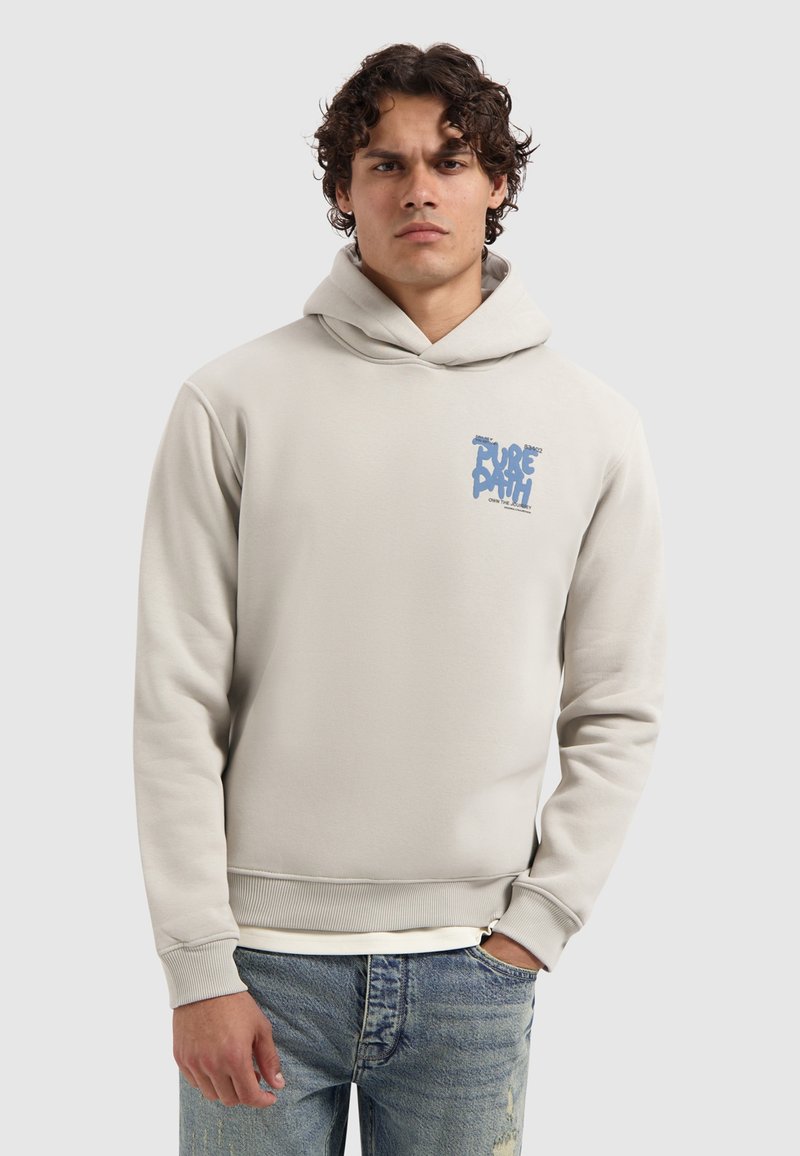 Mann in hellbeiger Kapuzenjacke mit blauer "PURE PATH"-Schrift auf der Brust, kombiniert mit hellblauen, verwaschenen Jeans, steht vor einem einfarbigen Hintergrund.