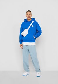 Blå hettegenser med frontlomme og grafikk, parret med lyseblå jeans og hvite joggesko. Tilbehør: hvit crossbody-veske.