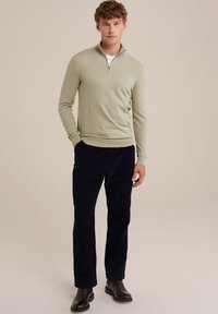 Šviesiai žalia užtraukiama megztinė, derinamas su tamsiai mėlynomis aksominėmis kelnėmis. Megztinis turi raštuotą apykaklę ir ilgas rankoves. Juodi odiniai batai užbaigia stilių.