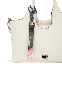 White textured leather tote bag with black accents, featuring a decorative tassel and a cartoon face emblem. Circular handles for carrying.