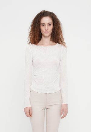 Haut blanc à manches longues avec motif cachemire, tissu transparent et détail froncé à la taille. L'encolure présente un bord festonné.