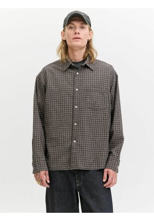 Young person with long blonde hair wearing a black baseball cap, brown checked button-up shirt, and dark jeans against a plain white background.