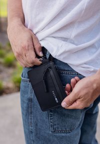 Black wallet made of textured material, featuring a zipper, a clip, and embossed branding. Positioned in a denim pocket, held by a hand.