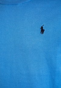 Blå stickad tröja med rund hals och ribbad textur, med en liten svart broderad logotyp på bröstet.