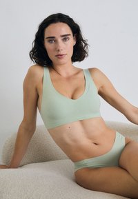 Model seated on a soft beige surface, wearing a light green bra and matching panties, with curly hair and a neutral expression.