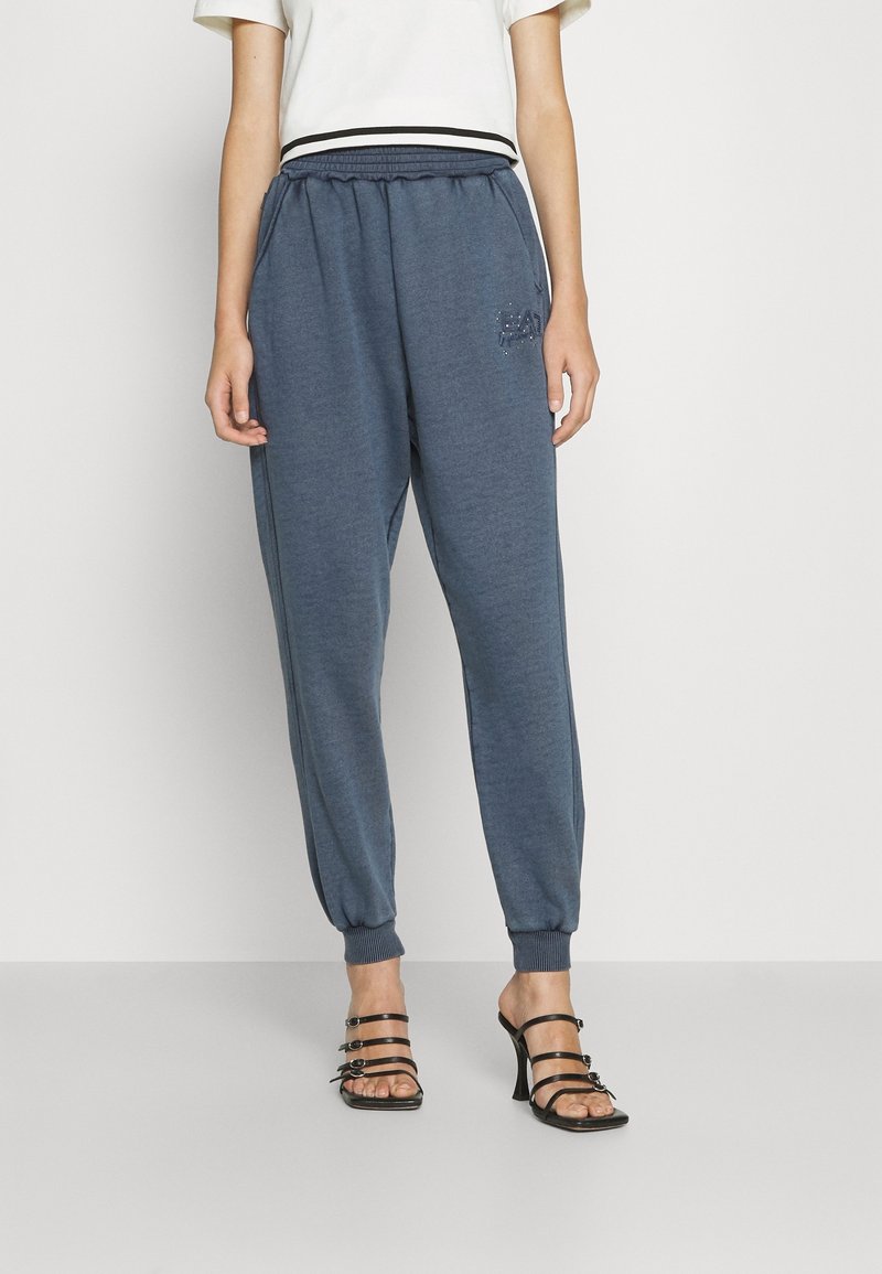 Dark blue sweatpants with an elastic waistband, ribbed cuffs, and a small logo detail on the side. Paired with strappy black heeled sandals.