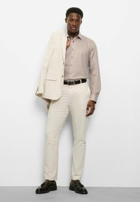 Heller Anzug mit einem cremefarbenen Blazer, einem gestreiften braun-weißen Hemd und hellen Hosen. Schwarze elegante Schuhe und ein schwarzer Gürtel vervollständigen den Look.