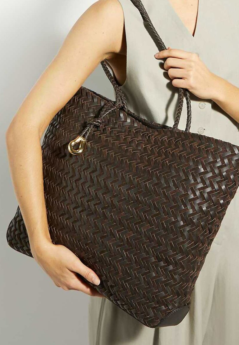 Woven tote bag in dark brown with a textured pattern, featuring braided straps and a gold accent. Generous, rectangular shape.