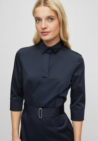 Sötétkék ingruha sima anyagból, galléros nyakkal, félhosszú ujjal és fekete övvel, amelynek téglalap alakú ezüst csatja van.