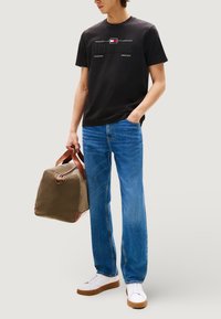 Maglietta nera in cotone con logo ricamato "HILFIGER", jeans in denim blu, sneakers bianche e borsa da viaggio verde oliva con dettagli in pelle marrone.