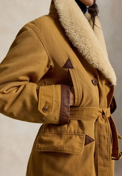 Καφέ παλτό με ζώνη, μεγάλο κρεμ shearling γιακά και δερμάτινη επένδυση στα μανίκια και στην τσέπη, που φοριέται από άτομο με το χέρι στην τσέπη.