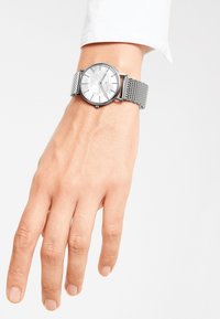 Silberne Mesh-Armbanduhr mit einem runden, weißen Zifferblatt, minimalistischen grauen Stundenmarkierungen und einem schlanken, polierten Gehäuserand.