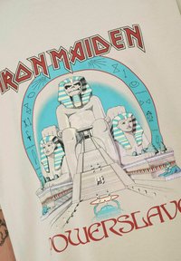  camiseta beige claro con un estampado gráfico de estatuas egipcias, rayas azules y blancas, y el texto rojo "Iron Maiden Powerslave".
