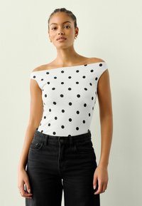 Biały top typu off-the-shoulder w czarne kropki, wykonany z elastycznego materiału. Połączony z czarnymi jeansami o wysokim stanie. Prosty design.