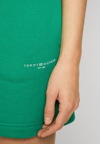 Zelený bavlněný jersey oděv s žebrovanou strukturou. Obsahuje bílé tištěné logo: "TOMMY HILFIGER EST. 1985" poblíž lemu.