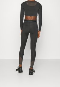 Musta urheiluasu, jossa on lyhythihainen toppi verkkoyksityiskohdilla ja korkeavyötäröiset legginsit. Molemmat ovat sileää, joustavaa materiaalia.