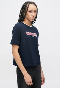 Tumesinist puuvillast t-särk, millel on valge ümargune kaelus ja esiküljel julge punane "tommy JEANS" logo, lõdvestunud lõige ja lühikesed varrukad.