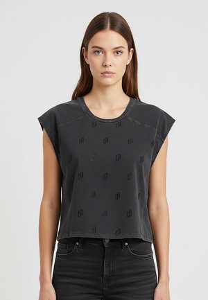 Giovane donna con capelli castani lisci indossa una canotta nera con un motivo sottile, abbinata a jeans neri a vita alta, in posizione neutra.