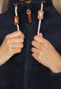 Veste en polaire bleu foncé avec un col haut, dotée de cordons tressés et d'accents en cuir marron au niveau du cou et de la fermeture éclair.