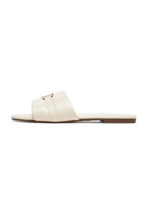 Flad slip-in sandal i cremefarve med bred rem og lille guldbeslag, vist fra siden på hvid baggrund.