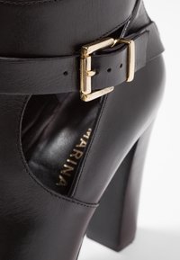 Zapato de tacón alto de cuero negro con un diseño recortado. Presenta una correa con hebilla dorada y sutilezas en los detalles de costura. Base de tacón plano.