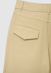 Beige Hosen mit strukturiertem Stoff, geradem Bein und einer einzelnen, schräg angesetzten Gesäßtasche. Der Bund ist mit Gürtelschlaufen versehen.