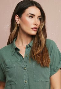 Camicia verde con bottoni, maniche corte, due tasche frontali e un tessuto morbido e texturizzato. Presenta un colletto classico e una leggera lucentezza.