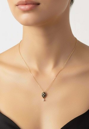Collier délicat en or avec un pendentif ballon à air chaud noir et or porté sur un cou et des épaules nus sur un fond clair.