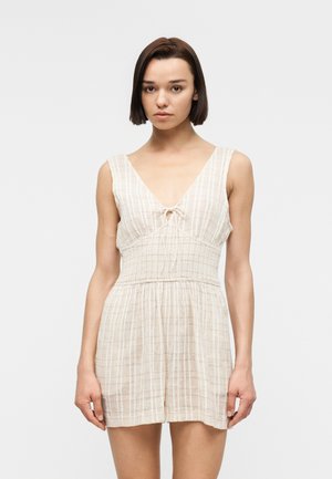 Jeune femme aux cheveux courts et foncés portant une combinaison-short sans manches à carreaux beige clair, avec un décolleté en V profond et une taille froncée.