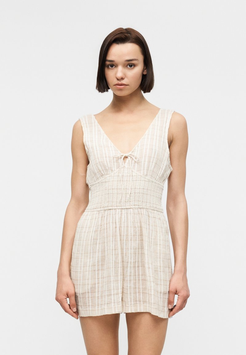 Jeune femme aux cheveux courts et foncés portant une combinaison-short sans manches à carreaux beige clair, avec un décolleté en V profond et une taille froncée.