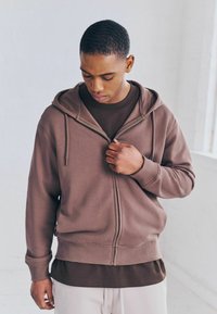 Sudadera con cremayera marrón de tela texturizada, con bolsillos frontales y cordones ajustables, superpuesta sobre una camiseta marrón más oscura.