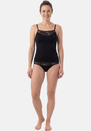 Frau steht barfuß da, trägt ein schwarzes Camisole mit Spitzenbesatz und passende schwarze Spitzenunterwäsche, lächelt in die Kamera.