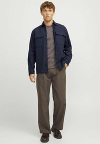 Giovane uomo con capelli ricci rossi che indossa una camicia blu scuro bottonata sopra una maglietta grigia, pantaloni marroni a pieghe e scarpe marroni scure, in una posa neutra.