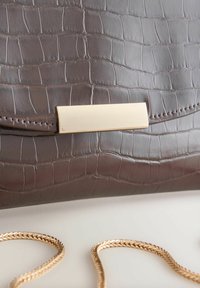 Braune Handtasche aus Kunstkrokodilleder mit glatter Textur, ausgestattet mit einem goldfarbenen Metallverschluss und einem geflochtenen Schulterriemen.