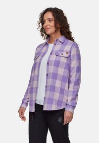 Camicia flanella a quadri viola e rosa chiaro con maniche lunghe, due tasche sul petto con bottone e chiusura frontale con bottoni, abbinata a pantaloni neri.