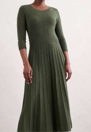 Frau trägt ein langes olivgrünes, geripptes Strickkleid mit Dreiviertelärmeln und tailliertem Oberteil vor einem schlichten Hintergrund.