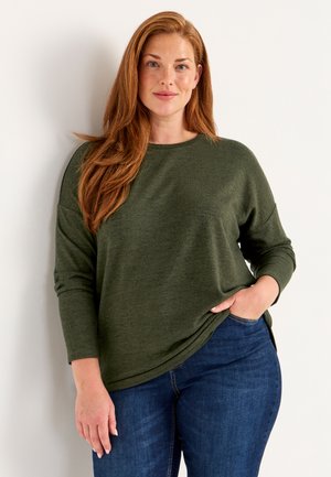 Olivgrünes, langärmliges Oberteil mit Rundhalsausschnitt und lockerer Passform, kombiniert mit dunkelblauen Jeans. Der Stoff wirkt weich mit einer subtilen Textur.