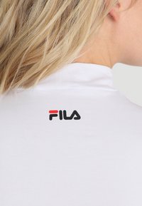 T-shirt polo blanc avec un col haut et un logo FILA noir et rouge au dos, avec une finition de tissu lisse et texturée.