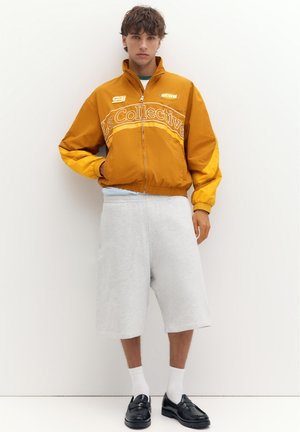 Jeune homme aux cheveux bouclés portant une veste zippée jaune moutarde, un short gris clair au genou, des chaussettes blanches et des mocassins noirs.