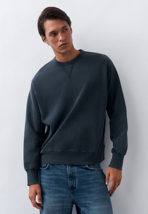 Jaunas vyras, vilkintis tamsiai pilką džemperį ir mėlynas džinsus, stovintis prie baltos sienos, žiūrintis į šoną neutraliu veidu.