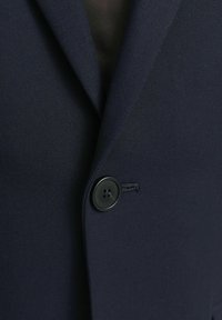 Tumesinine bleiser, millel on tekstureeritud kangas, struktureeritud klapid ja üks must nupp nelja auguga. Üksikasjalik õmblus on nähtav.