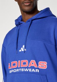 Modra Adidas kapuca z velikim rdečim logotipom "ADIDAS SPORTSWEAR", belim trikotnim emblemom in kapuco s vrvico. Mehka tkanina in sproščen kroj.