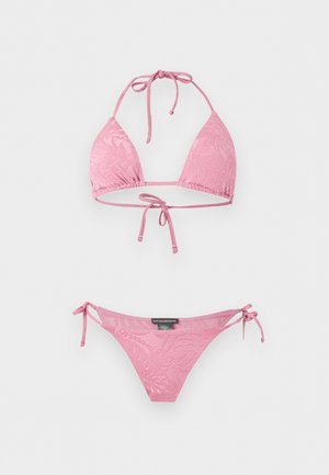 Γυναίκα που στέκεται ξυπόλητη φορώντας ροζ μπικίνι με σχέδια και δέσιμο στα πλάγια, πάνω σε απλό λευκό φόντο, με το χέρι στη μέση.