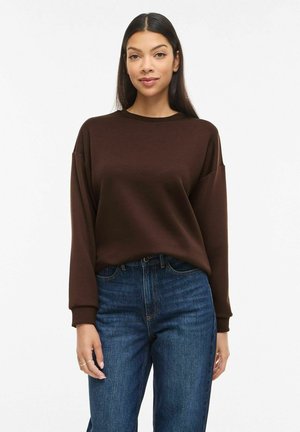 Femeie cu păr drept purtând un pulover cu mâneci lungi de culoare maro închis, băgat în blugi cu talie înaltă, albastri, stând în fața unui fundal alb.