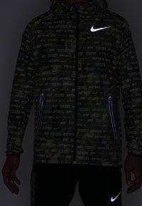 Zwarte Nike-jack met een donker patroon en herhaalde "Just Do It" tekst, wit Nike-logo op de borst en met rits afgesloten zijzakken, omrand door blauwe bies.