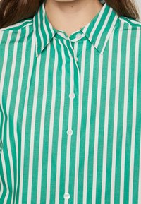Camicia a righe verdi e bianche con colletto classico, chiusura frontale con bottoni e tessuto leggero, caratterizzata da righe verticali e una texture semi-trasparente.