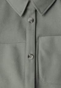 Szara koszula z mieszanki wełny z kołnierzykiem, zapinana na dwa jasnoszare guziki z przodu oraz z kieszonką na lewym piersi. Miękka faktura na całej powierzchni.